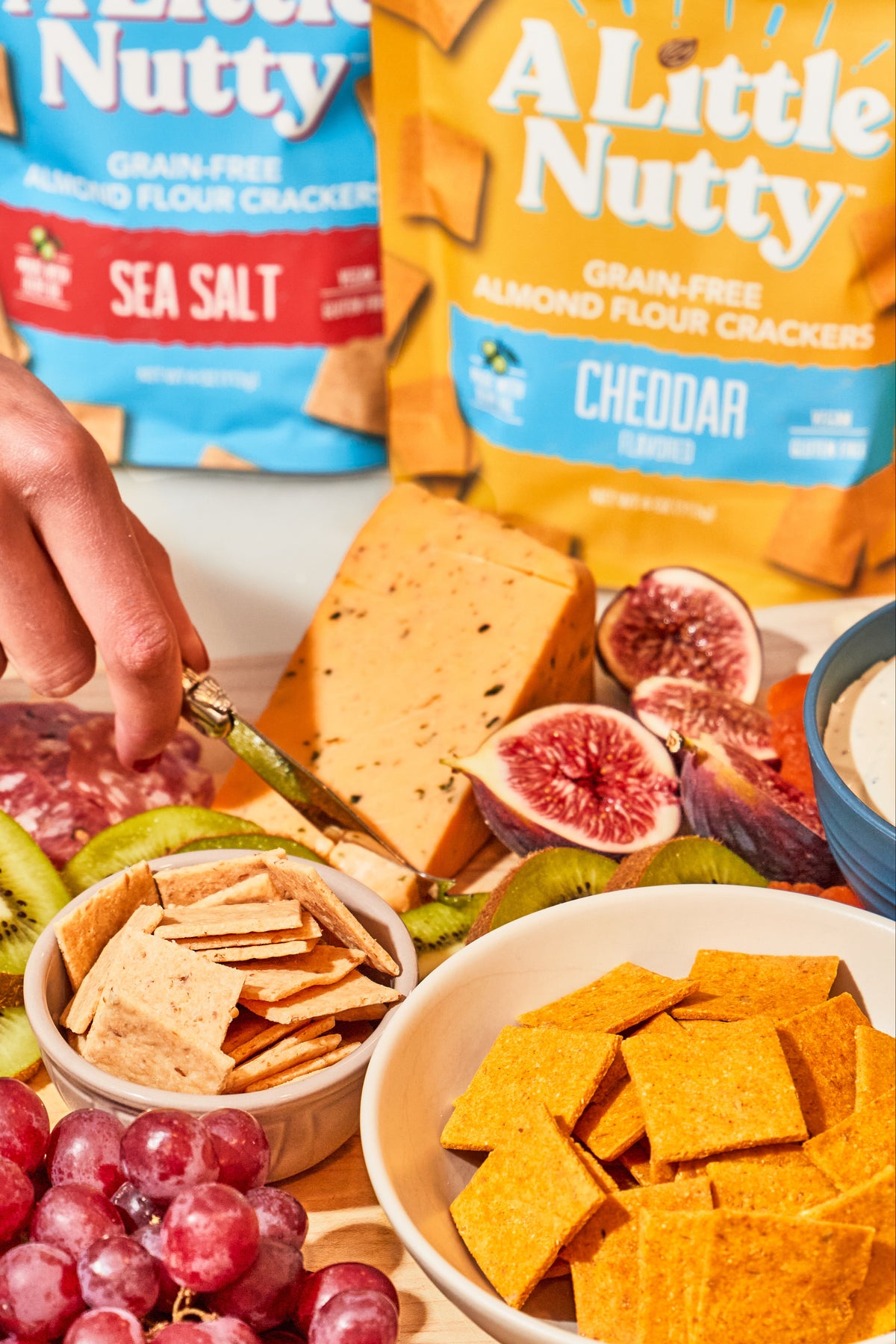 Table with crackers, fruits, and a hand reaching for a cracker, with 'A Little Nutty' packaging in the background.
