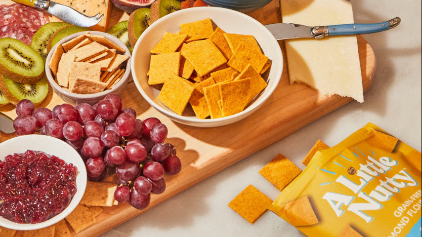 Assorted snacks including cheese, crackers, and fruits on a wooden board with Little Nutty packaging.